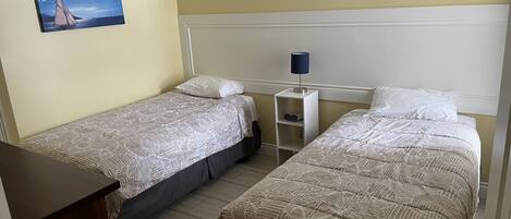 A hotel room with two single beds, a bedside table with a lamp, a small shelf, and a painting on the wall.