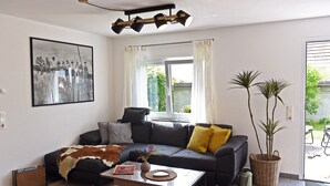 Una sala de estar con un sofá, una mesa de centro, un cuadro enmarcado, una ventana con cortinas y una planta en maceta.