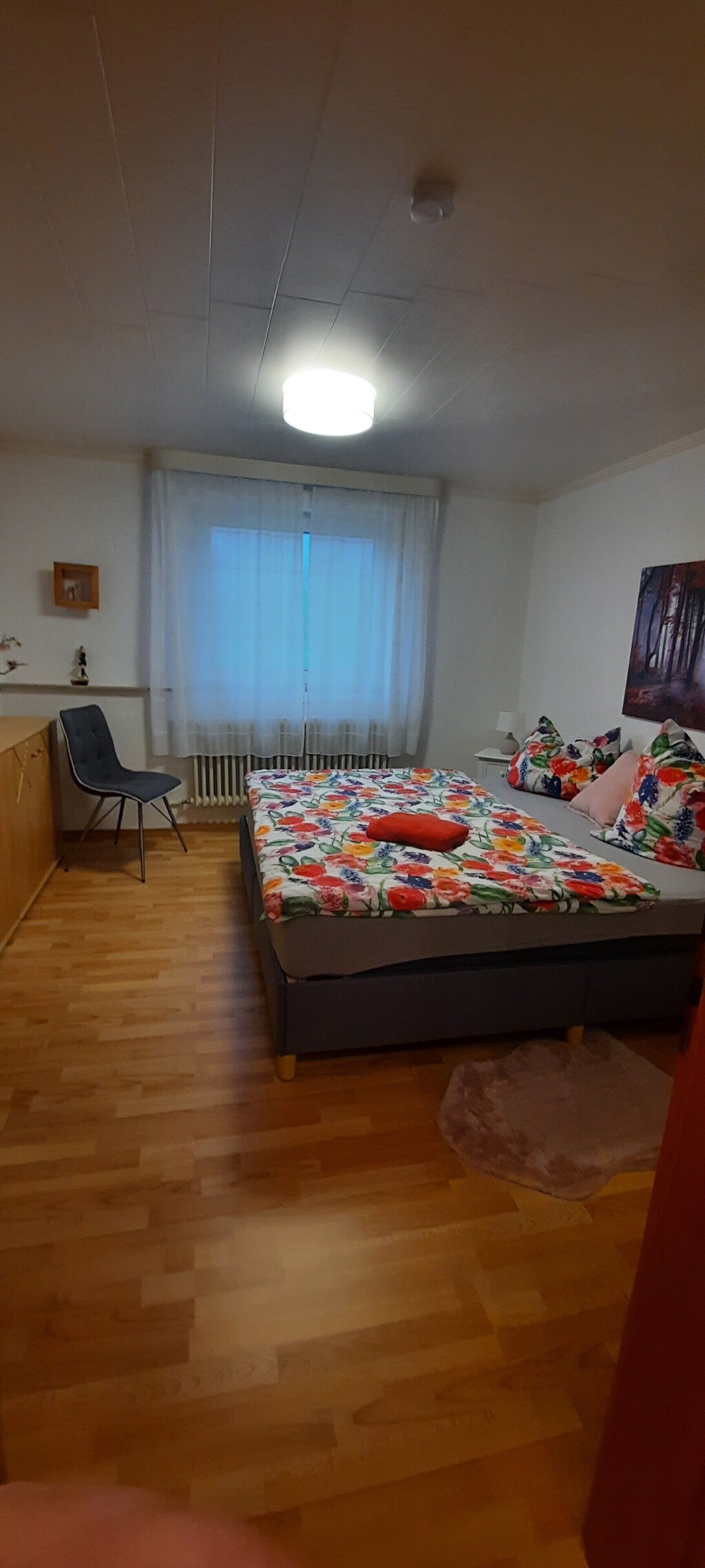 Makuuhuoneessa on sänky, tuoli, patteri ja ikkuna, jossa on verhot.
