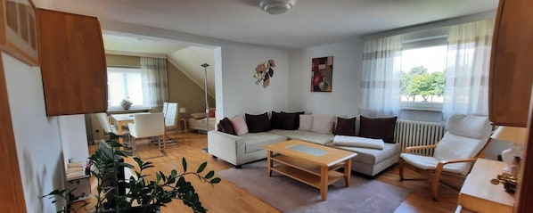 Un salon avec un canapé, une table basse et des fauteuils, un coin repas avec une table et des chaises, et un escalier menant à une autre pièce.