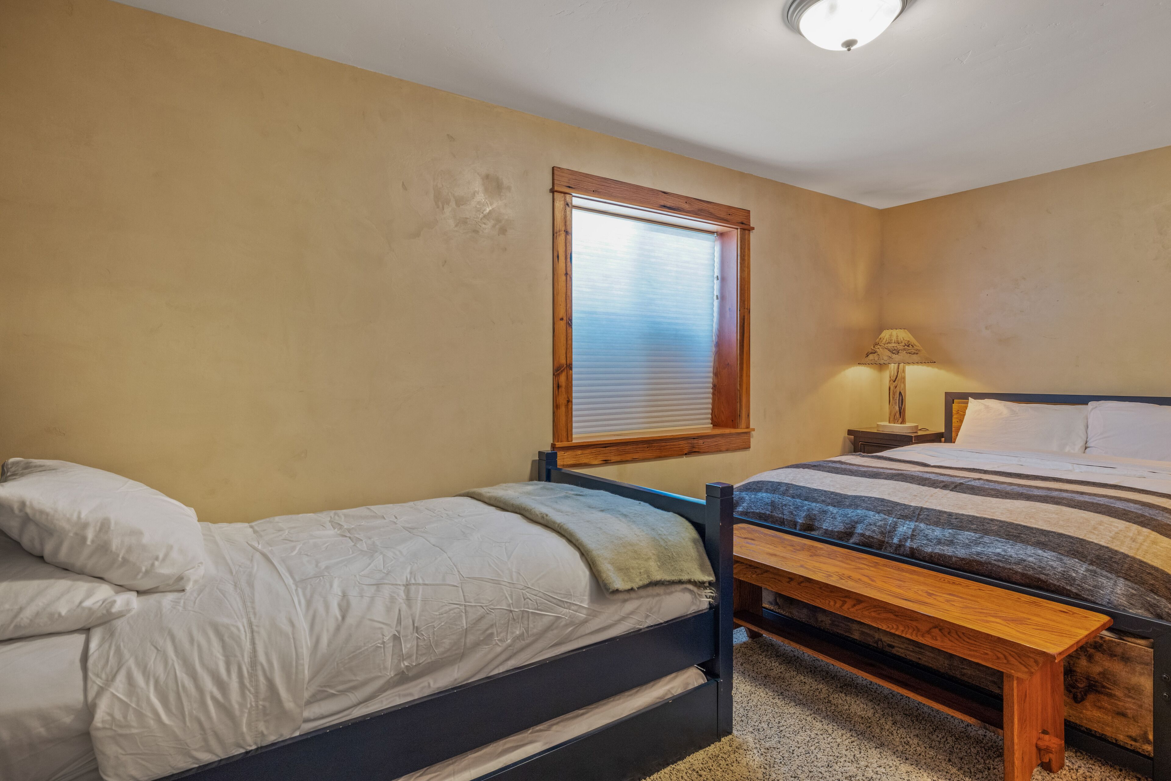 A bedroom with two single beds, a window with blinds, a bedside table with a lamp, and a carpeted floor.