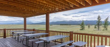 A covered outdoor area with picnic tables and benches, overlooking a grassy field and mountains.