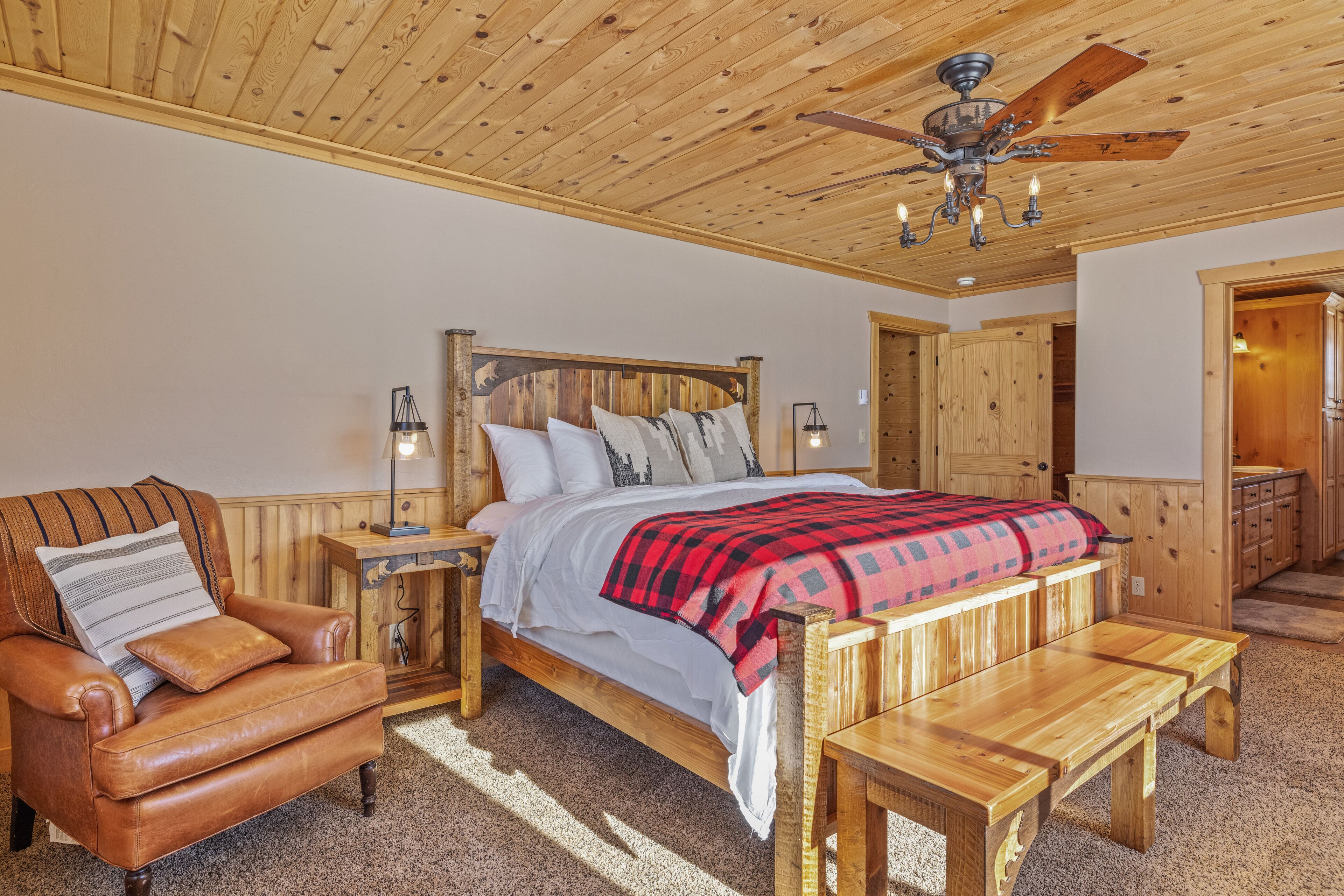 Ein Schlafzimmer mit Holzboden, einem Bett mit rot-schwarz kariertem Überwurf, einem Ledersessel, einer Holzbank und einem Nachttisch mit Lampe.