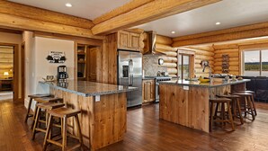 A spacious kitchen with wooden cabinetry, a large island with stools, a stainless steel refrigerator, and a view of the outdoors through a window. - Stone Skipper Lodge (Island Park)