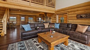 A spacious log cabin interior with a large L-shaped sectional sofa, a wooden coffee table, and a dining area with a wooden table and chairs. - Stone Skipper Lodge (Island Park)