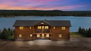A log cabin with a front porch and multiple windows, situated by a lake with a forested backdrop. - Stone Skipper Lodge (Island Park)