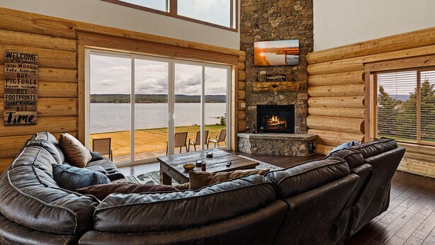 A log cabin interior with a leather sofa, a stone fireplace, and a large sliding glass door leading to a lakeside view. - Stone Skipper Lodge (Island Park)