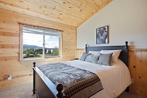 A bedroom with a wooden ceiling, a large window with a view of mountains, a bed with a patterned blanket, and a framed picture on the wall. - Henrys Hawk (Island Park)