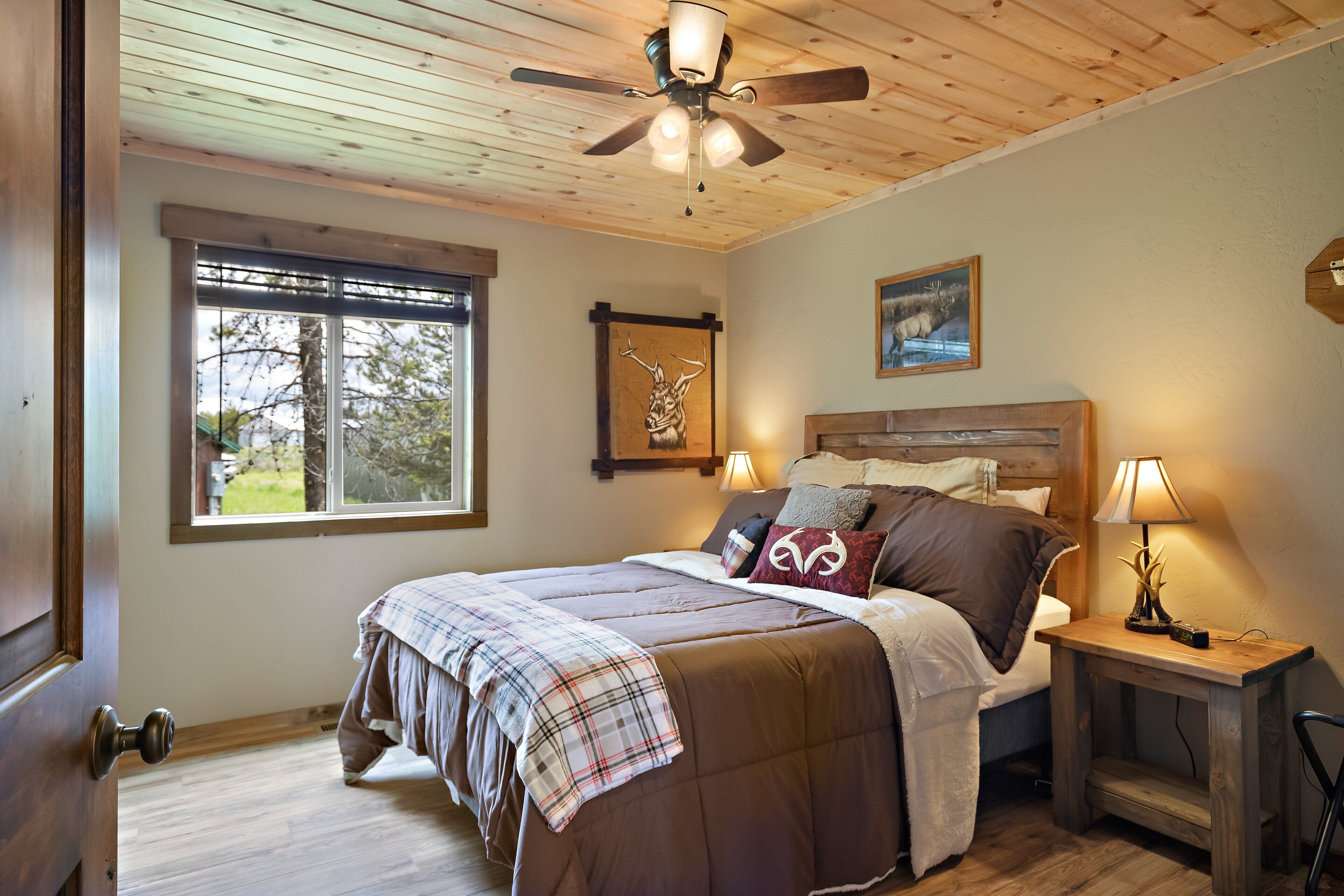 A bedroom with a wooden ceiling, a large bed with a plaid blanket, a nightstand with a lamp, and a window with a view of trees.
