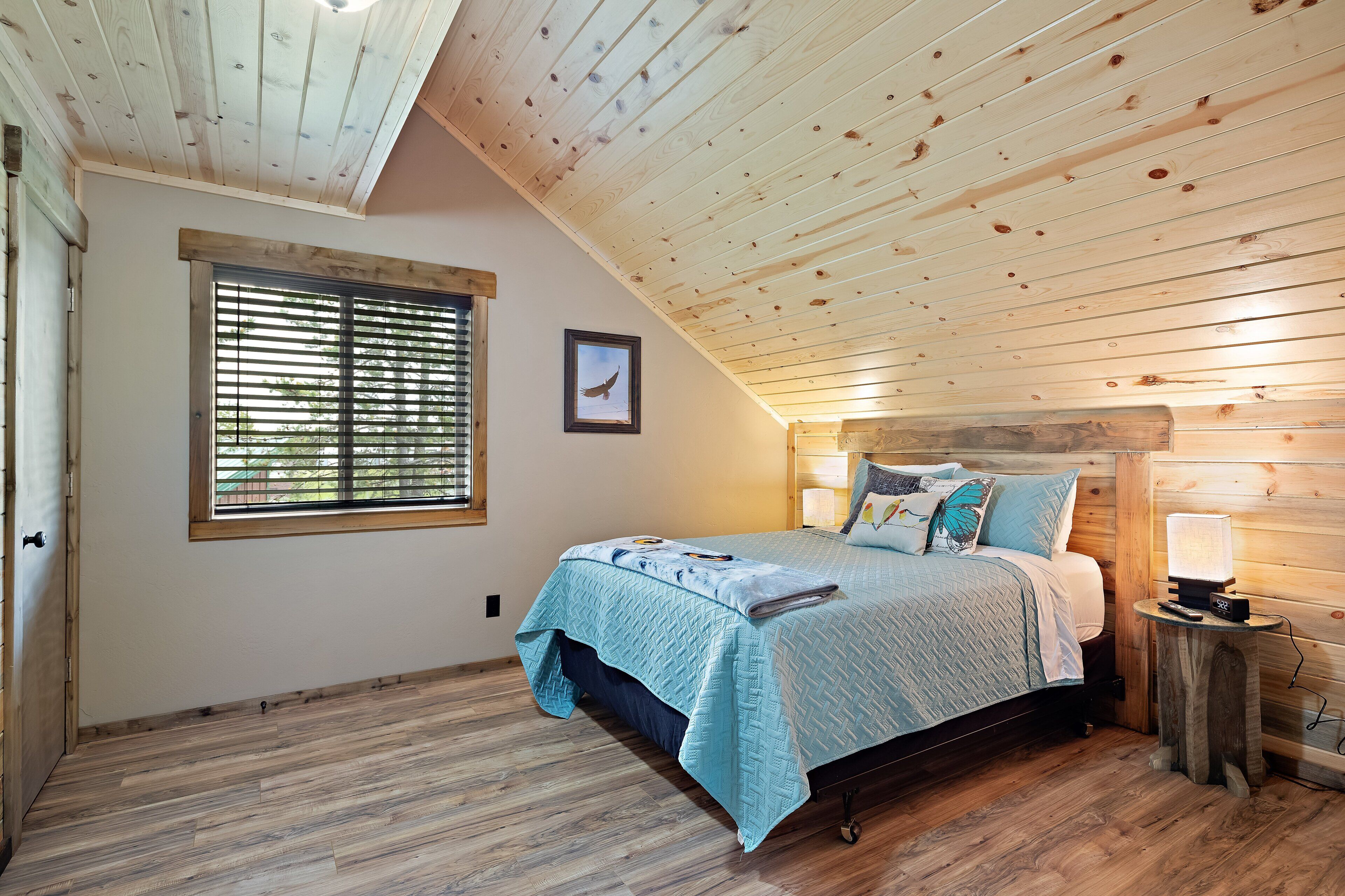 Ein Schlafzimmer mit Holzbalkendecke, einem Bett mit blauer Steudecke, einem Nachttisch mit Lampe und einem Fenster mit Jalousien.