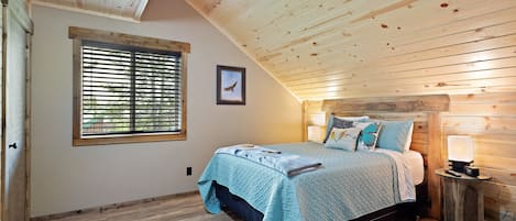 Ein Schlafzimmer mit Holzbalkendecke, einem Bett mit blauer Steudecke, einem Nachttisch mit Lampe und einem Fenster mit Jalousien.