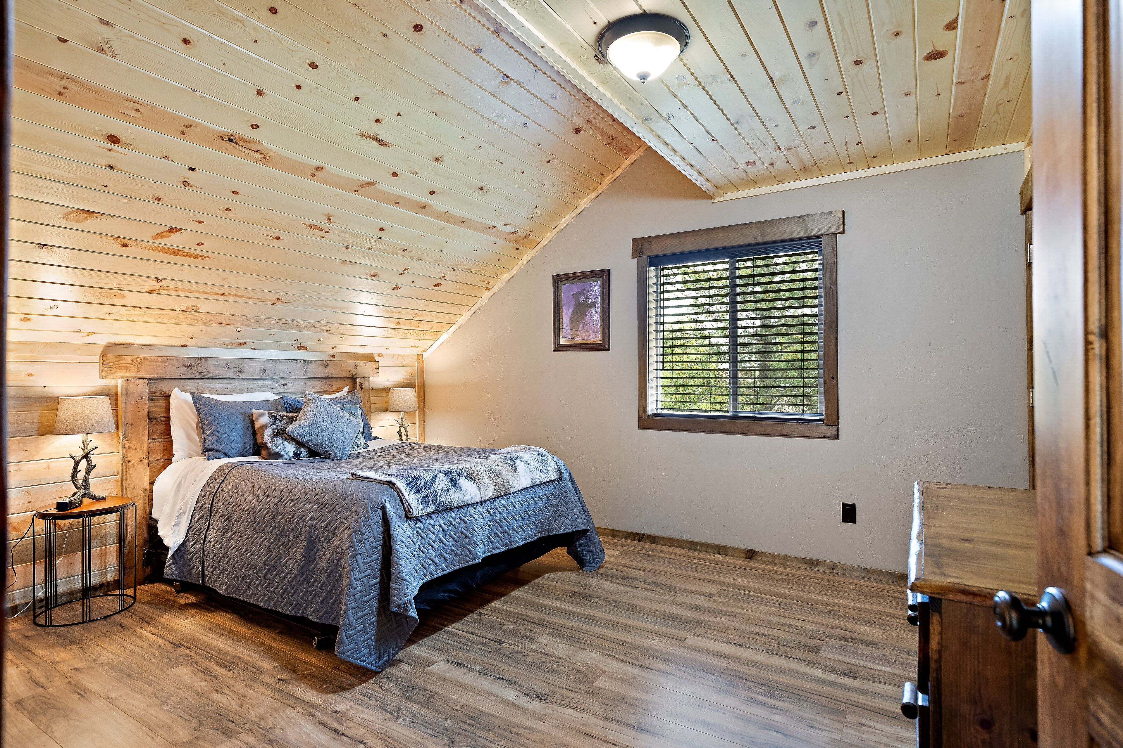 A bedroom with a sloped ceiling, wooden flooring, a bed with a patterned bedspread, a bedside table with a lamp, and a window with blinds.