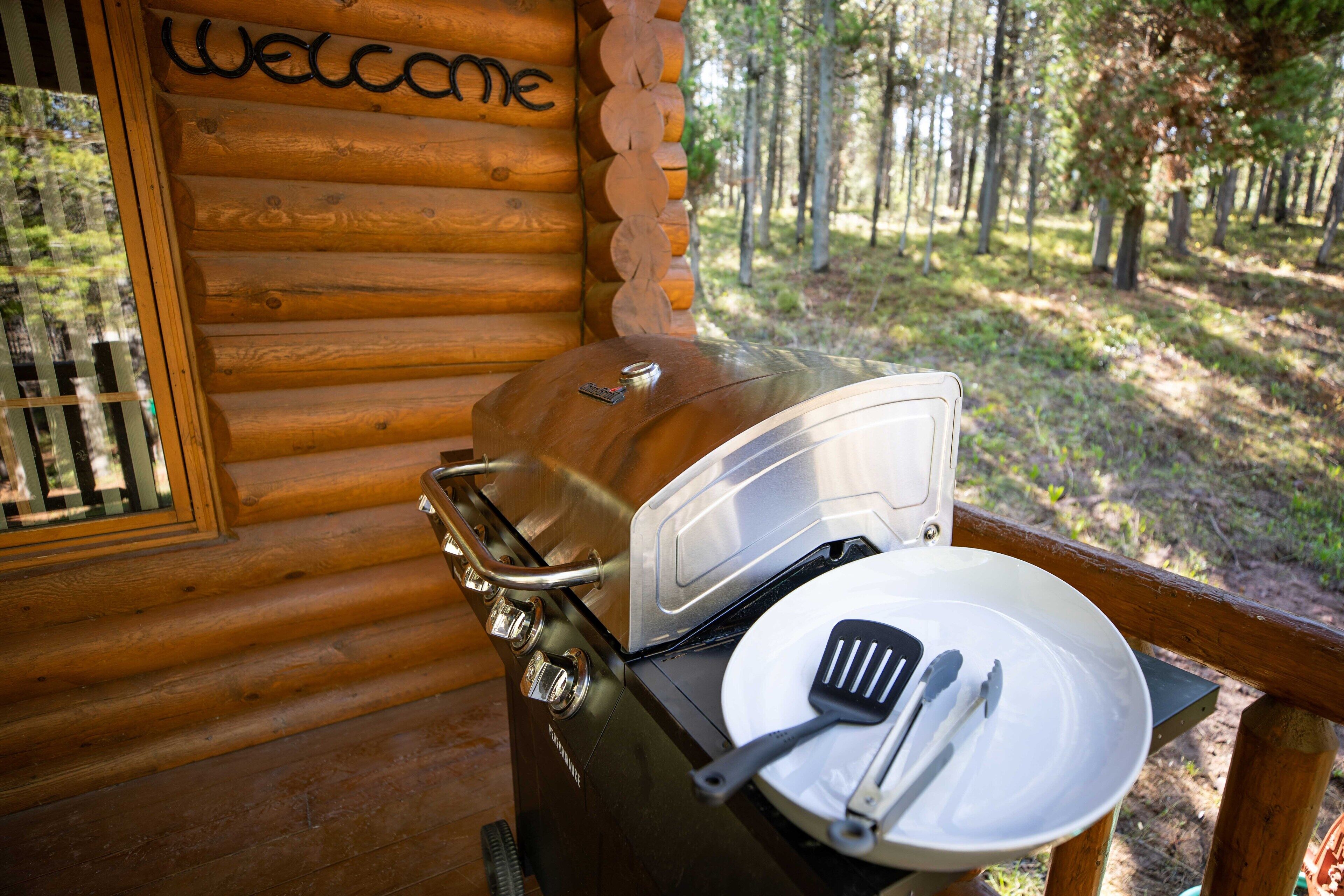 A wooden cabin with a 'Welcome' sign, a grill, and a plate with utensils on a table.