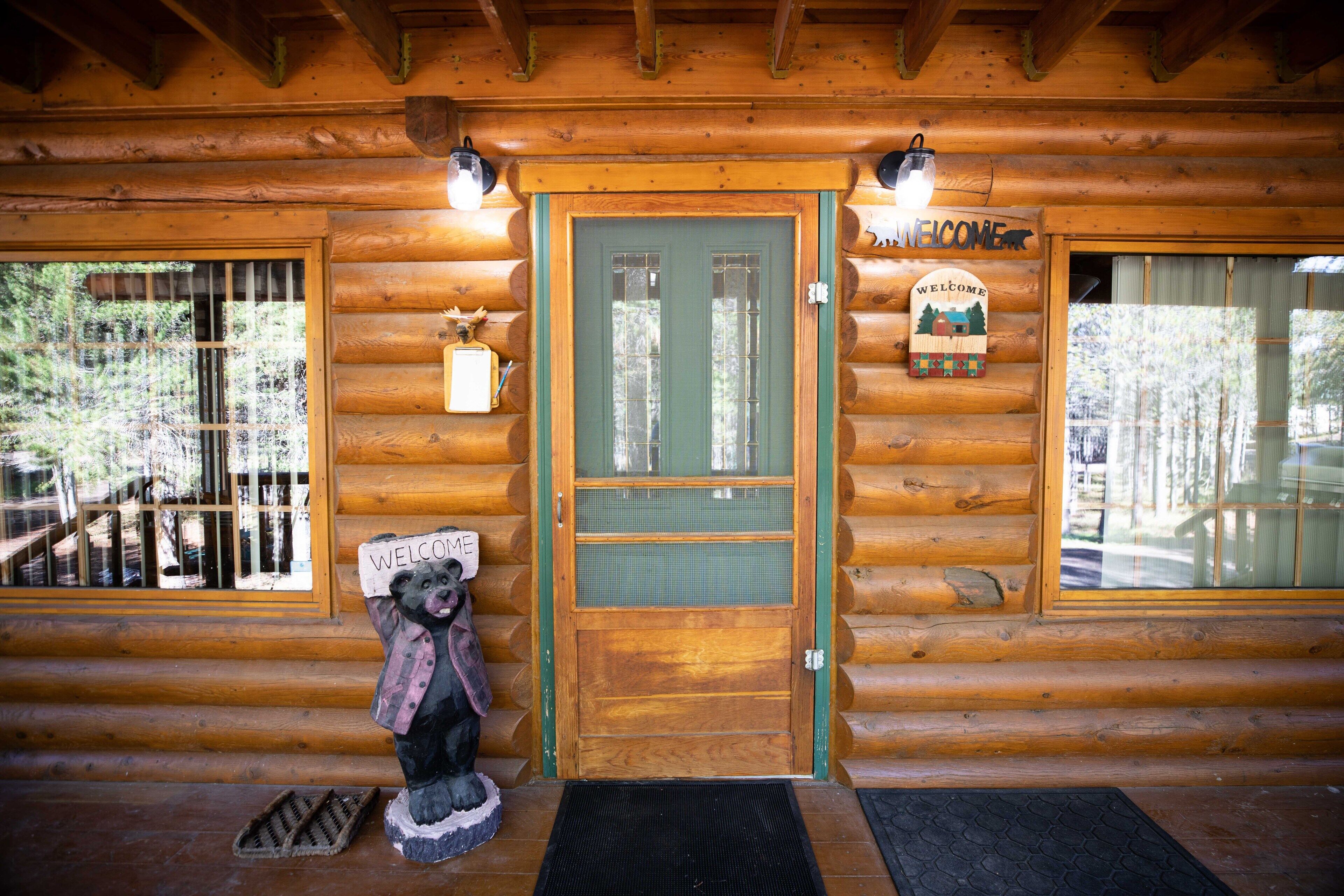 Une cabane en bois avec une porte verte, une statue d’ours tenant une pancarte et une enseigne « Bienvenue » au-dessus de la porte.