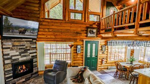Ein Holzhaus-Interieur mit Kamin aus Stein, einem an der Wand befestigten Fernseher, einer gemütlichen Sitzecke mit Sofa und Sesseln sowie einem Esstisch mit Stühlen.