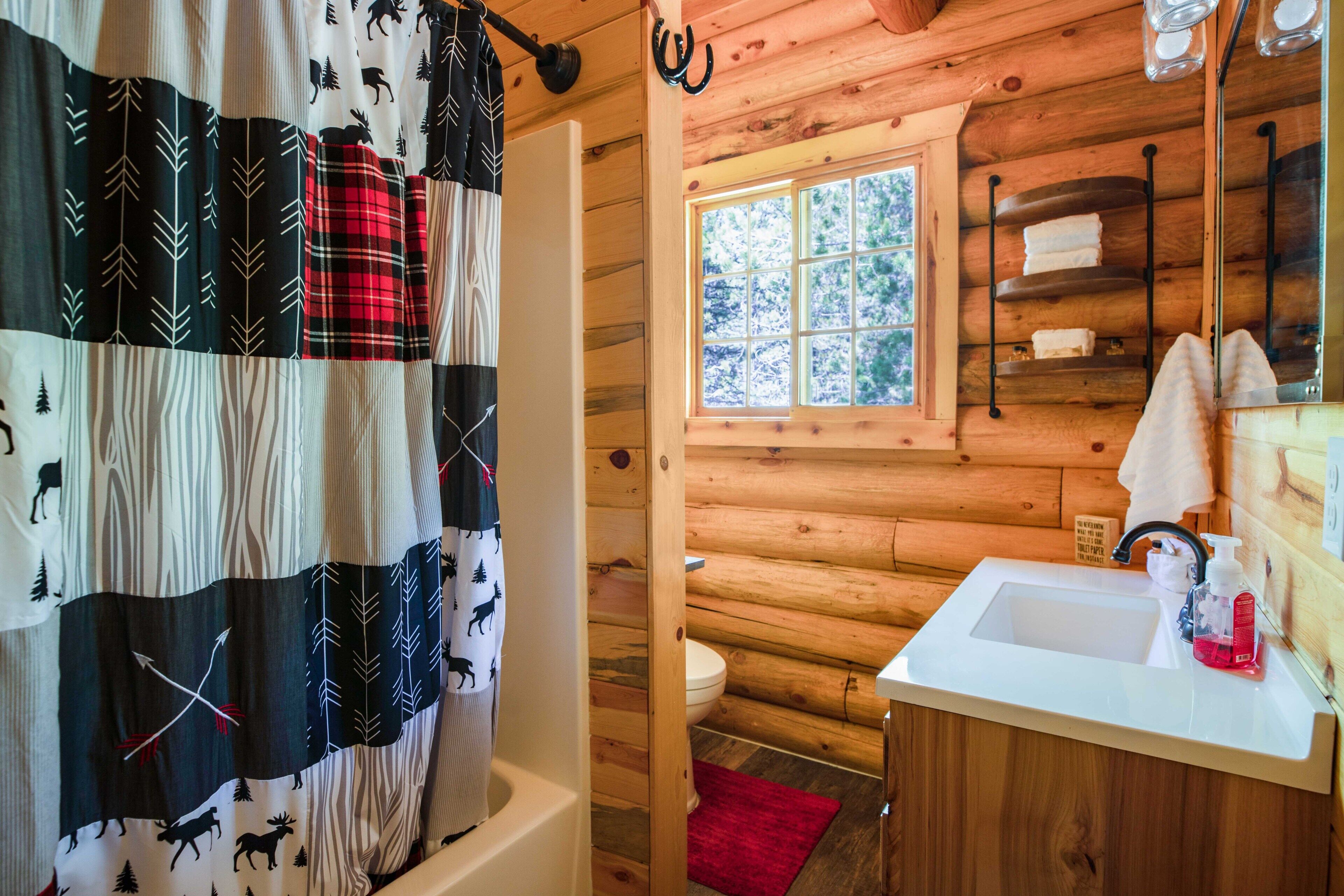 A wooden cabin bathroom with a shower curtain featuring animal prints, a white sink with a red mat, and a window with a view of trees.