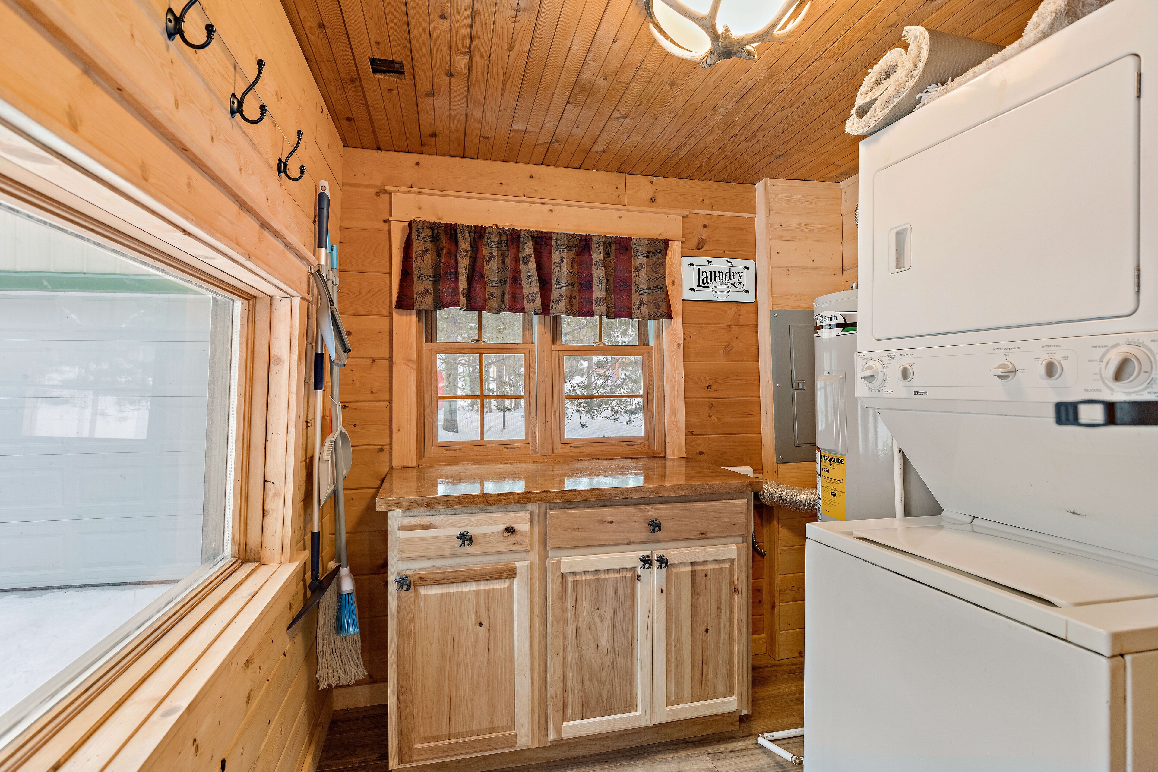 A wooden cabin interior with a kitchen area, including a wooden cabinet with drawers, a window with a view of a snowy landscape, and a washing machine.