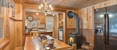 A wooden cabin interior with a dining table, chairs, a chandelier, a large refrigerator, and a wood-burning stove.