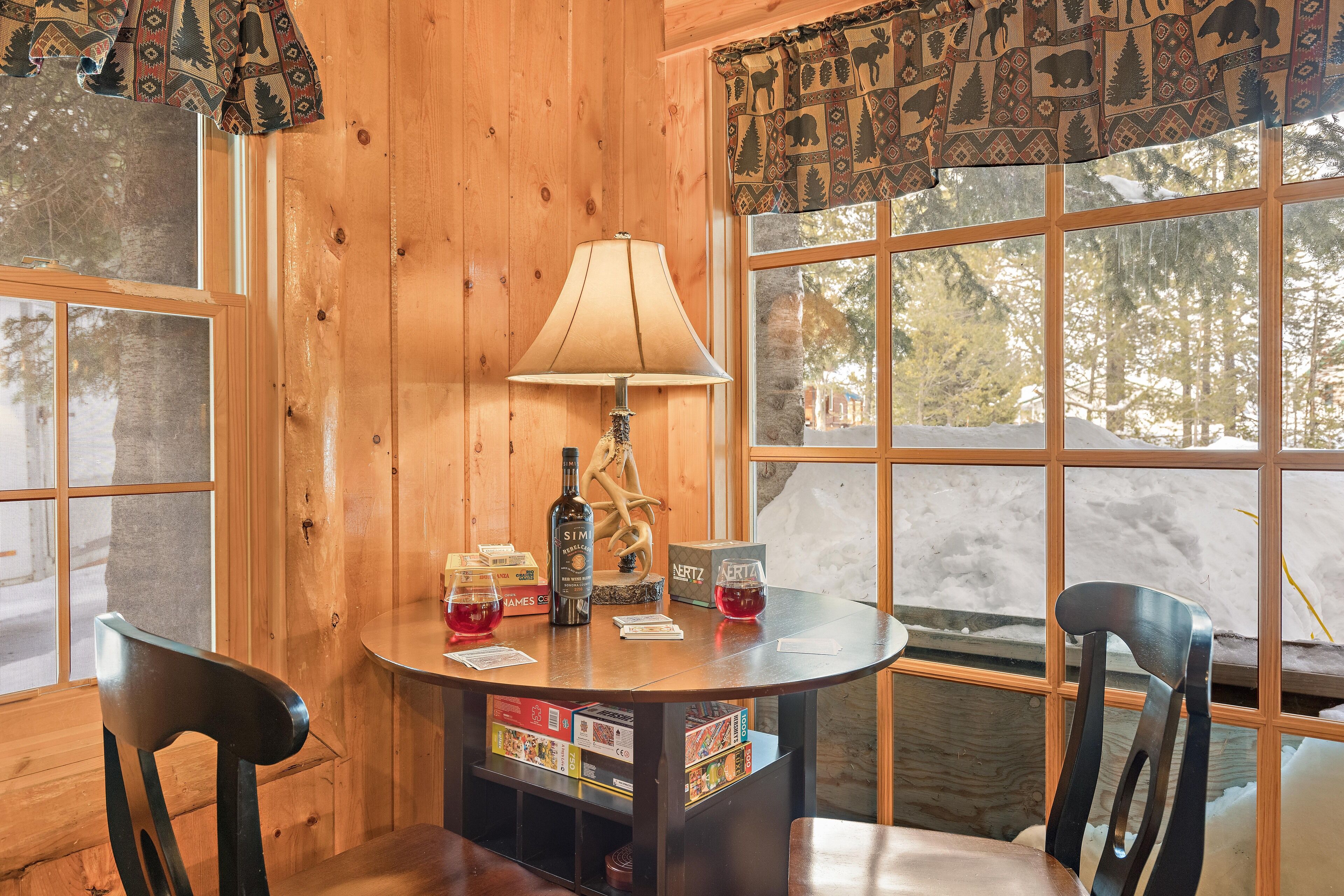 A wooden interior with a round table, chairs, a lamp, and a view of a snowy landscape through large windows.