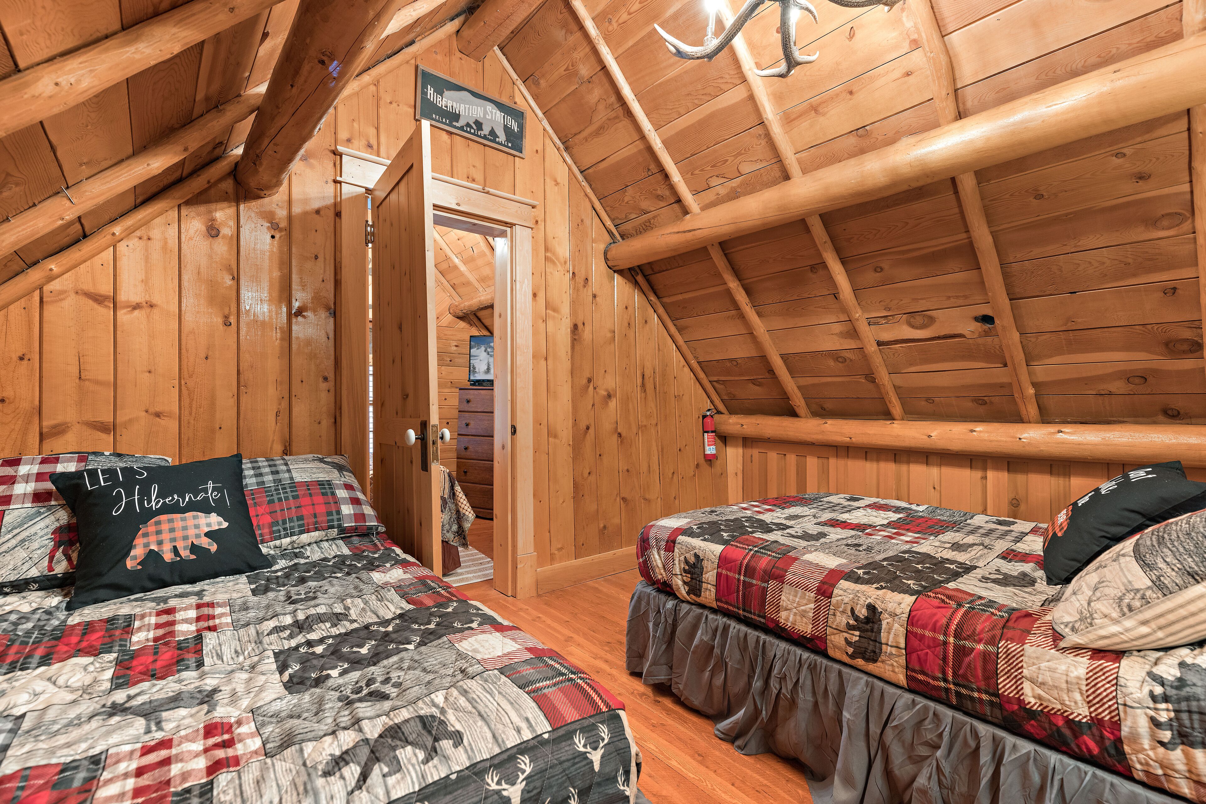 A cozy log cabin bedroom with two beds, a wooden ceiling, and a door leading to a bathroom.