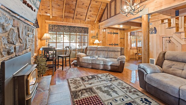 A cozy cabin interior with a stone fireplace, a plush sofa, a dining area, and a staircase leading to an upper level. - Double Loop (Island Park)