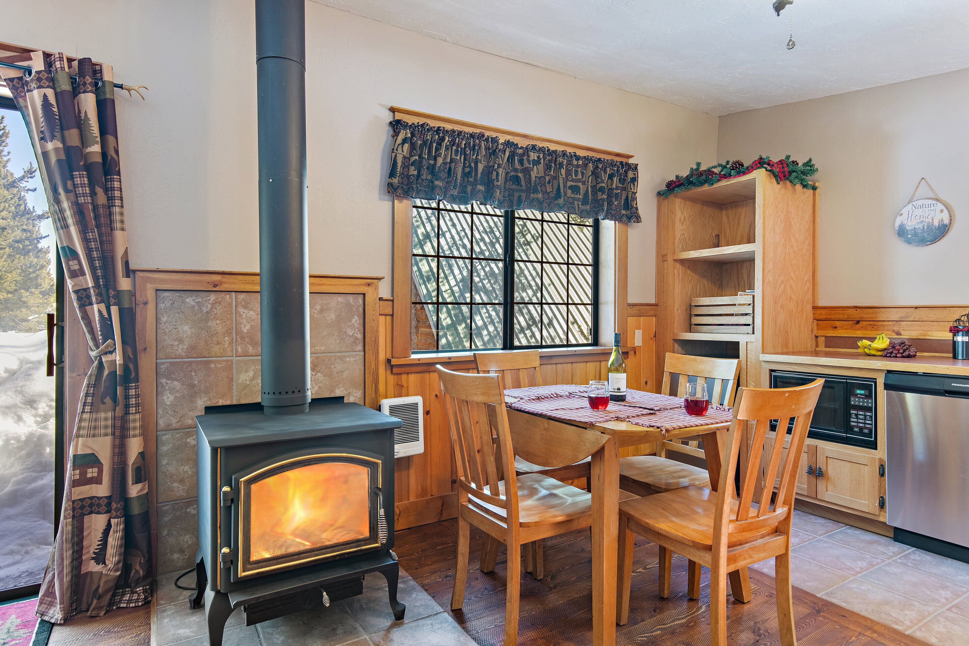 Una cocina acogedora con estufa de leña, sillas de madera, una mesa de comedor y un ventanal con vistas a árboles.