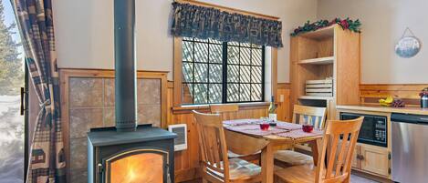 Una cocina acogedora con estufa de leña, sillas de madera, una mesa de comedor y un ventanal con vistas a árboles.