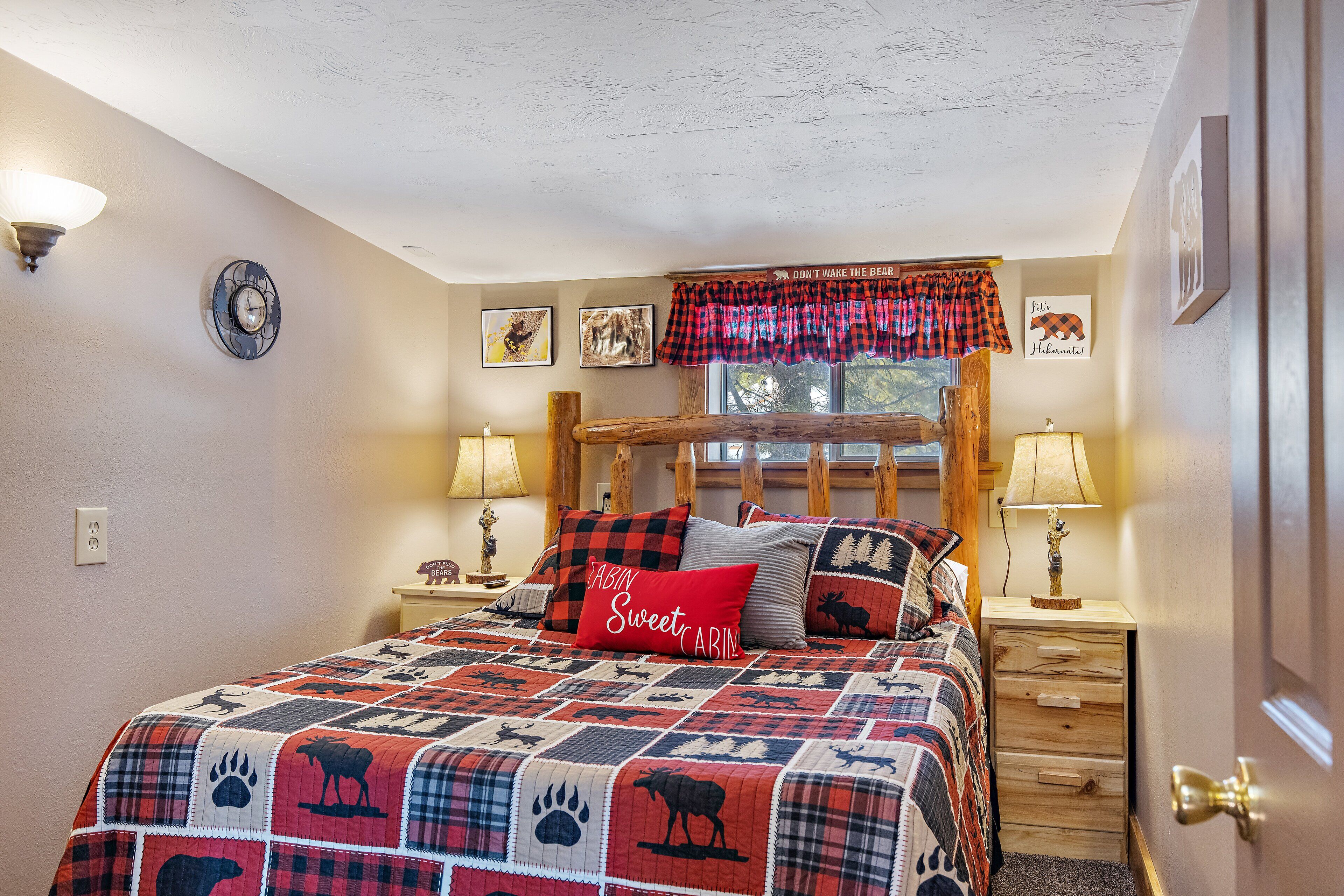 A bedroom with a bed, two bedside tables, a nightstand, a window with a plaid valance, and wall-mounted artwork.