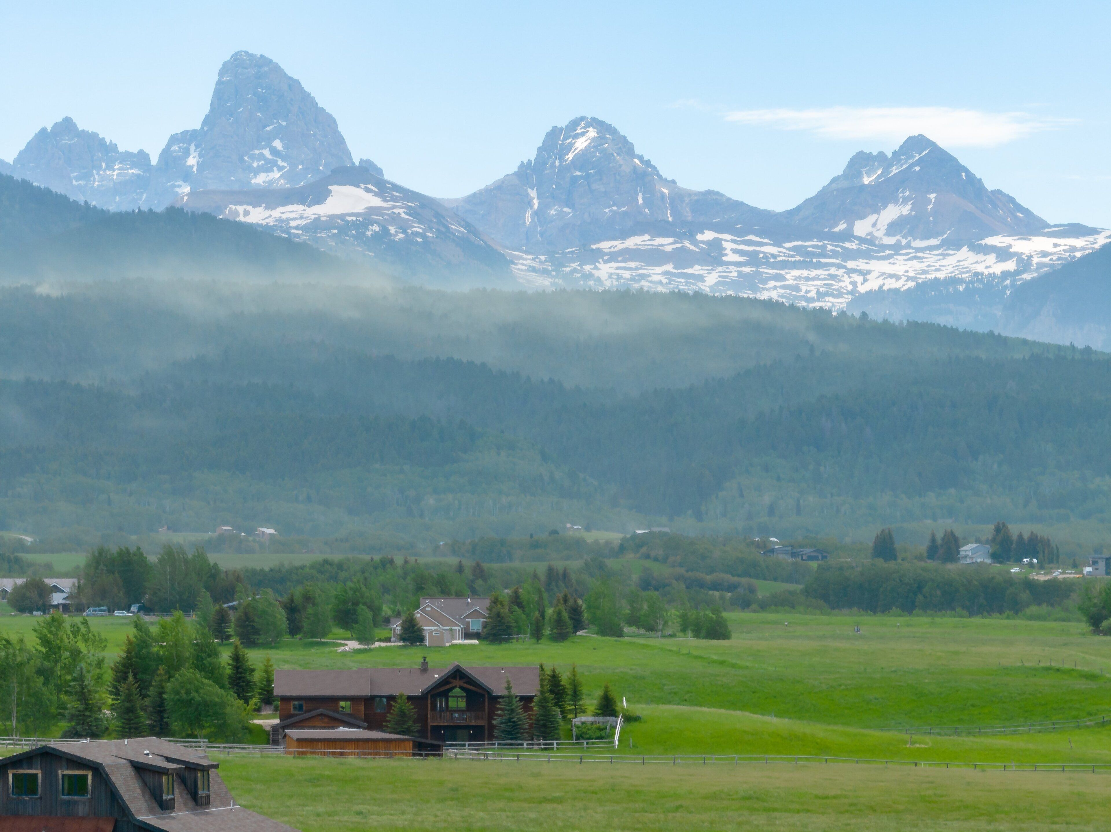 A rural landscape with a house surrounded by green fields and mountains in the background.