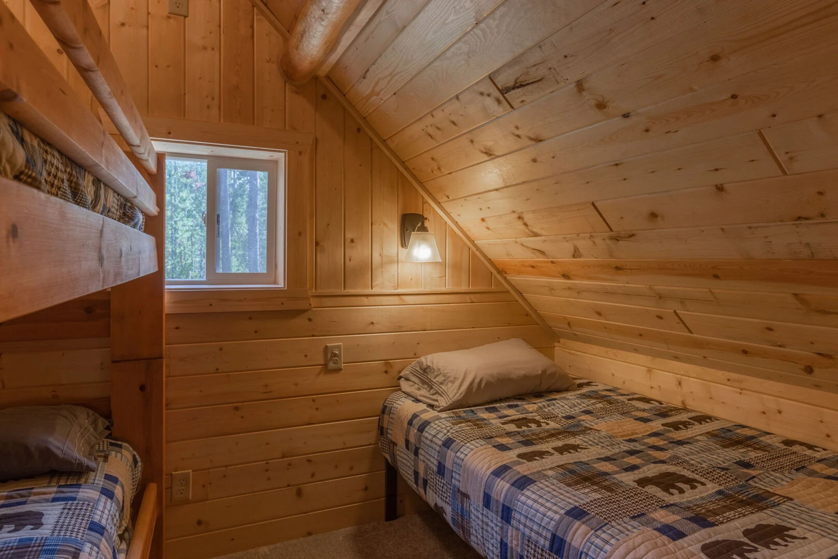 Acogedora habitación tipo cabaña con techo inclinado, paredes de madera, una ventana, una cama con colcha y una lámpara fijada a la pared.