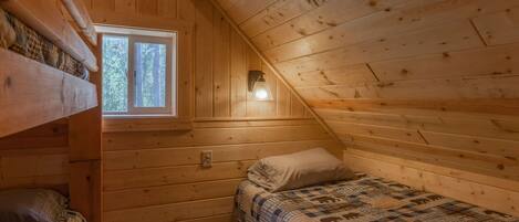 Acogedora habitación tipo cabaña con techo inclinado, paredes de madera, una ventana, una cama con colcha y una lámpara fijada a la pared.