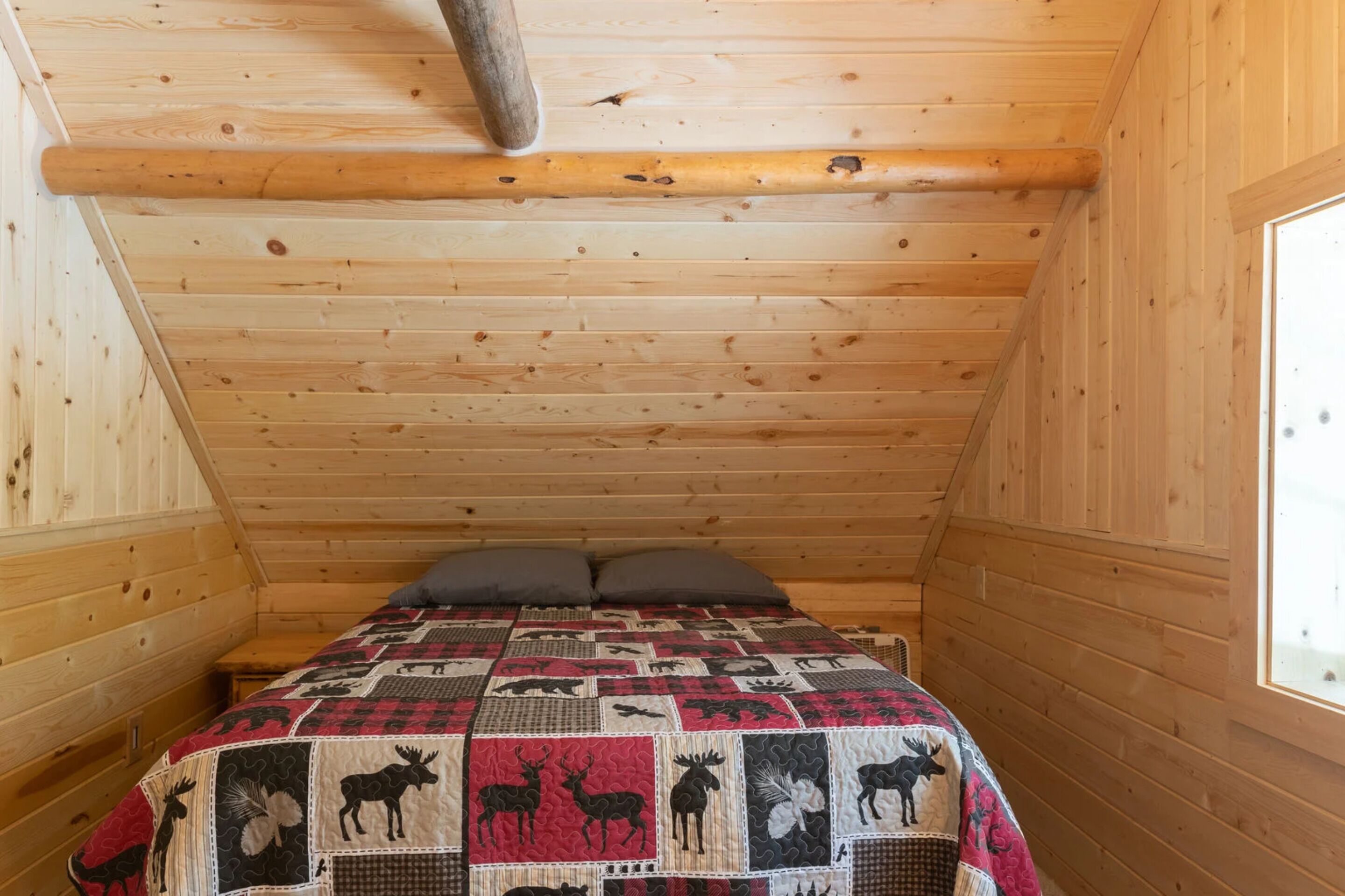 A cozy wooden cabin bedroom with a quilted bedspread featuring animal prints, a window on the right, and a wooden ceiling beam.