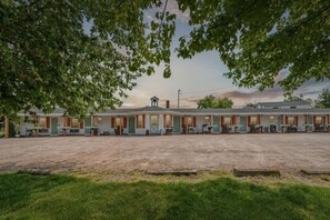 Ein Motel mit einem zentralen Gebäude und angebauten Zimmern, einem Parkplatz mit Kiesbelag und Bäumen, die das Bild säumen.