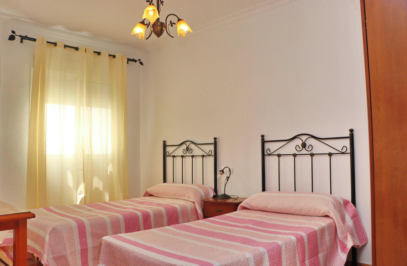 A bedroom with two single beds, a nightstand with a lamp, a chandelier, and a window with curtains.