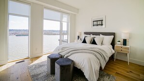A bedroom with a large bed, a nightstand, a lamp, and a view of a body of water through large windows. - Delux River View Home 2beds (New Westminster)