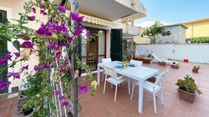 Patio esterno con tavolo e sedie bianche, circondato da piante in vaso e una pianta rampicante con fiori viola.