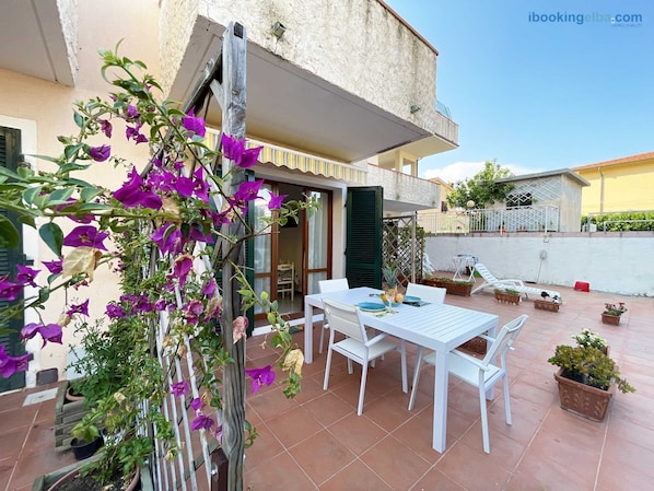 An outdoor patio with a white table and chairs, surrounded by potted plants and a climbing vine with purple flowers.