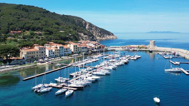 Una marina con numerose imbarcazioni ormeggiate, una cittadina costiera con edifici e una collina ricoperta di vegetazione.