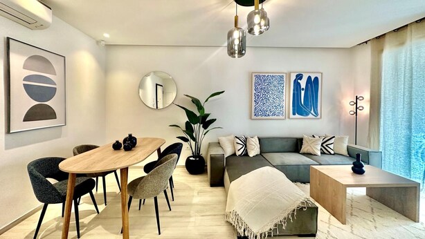 A modern living room with a dining area, featuring a wooden dining table, chairs, a sofa, and a coffee table. There are two wall-mounted light fixtures and a floor lamp. - A12 Stylish and Modern 2bds in La Marsa (La Marsa)