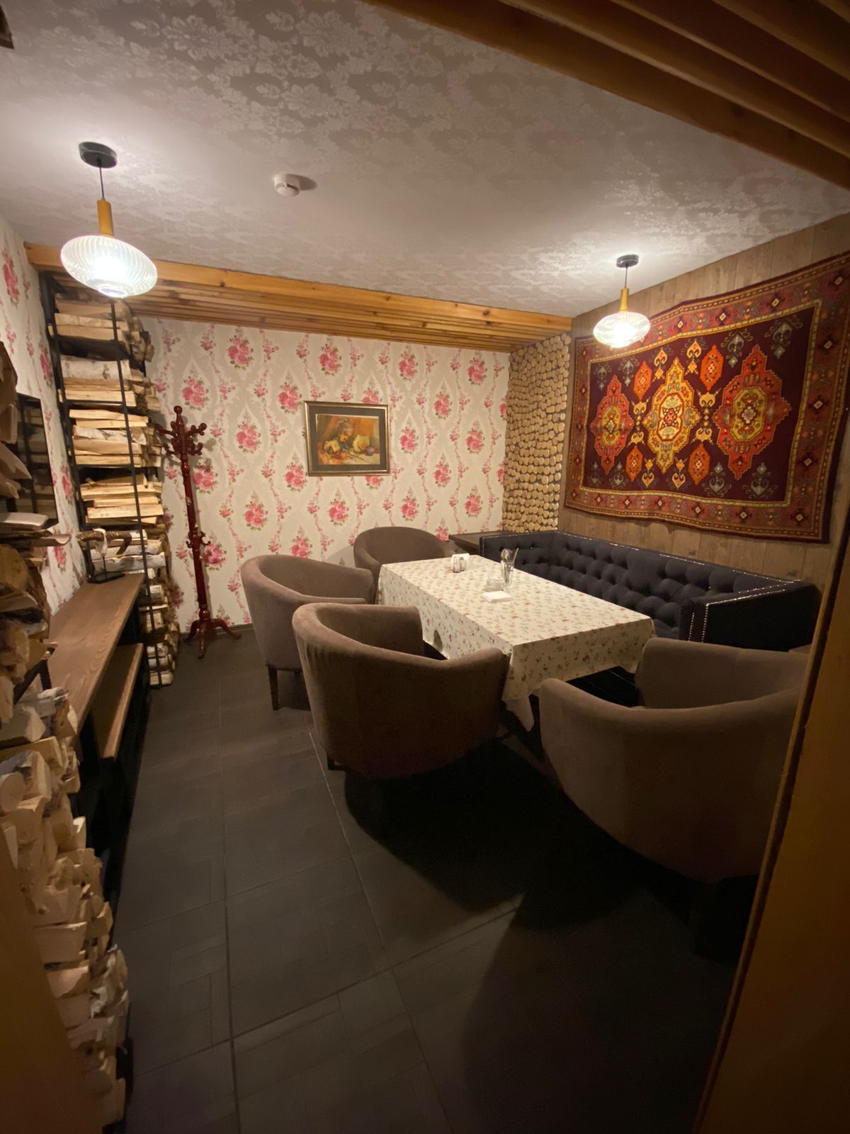 Un coin repas chaleureux avec un banc en bois, une table recouverte d’une nappe à motifs floraux et une tapisserie ornementale accrochée au mur.