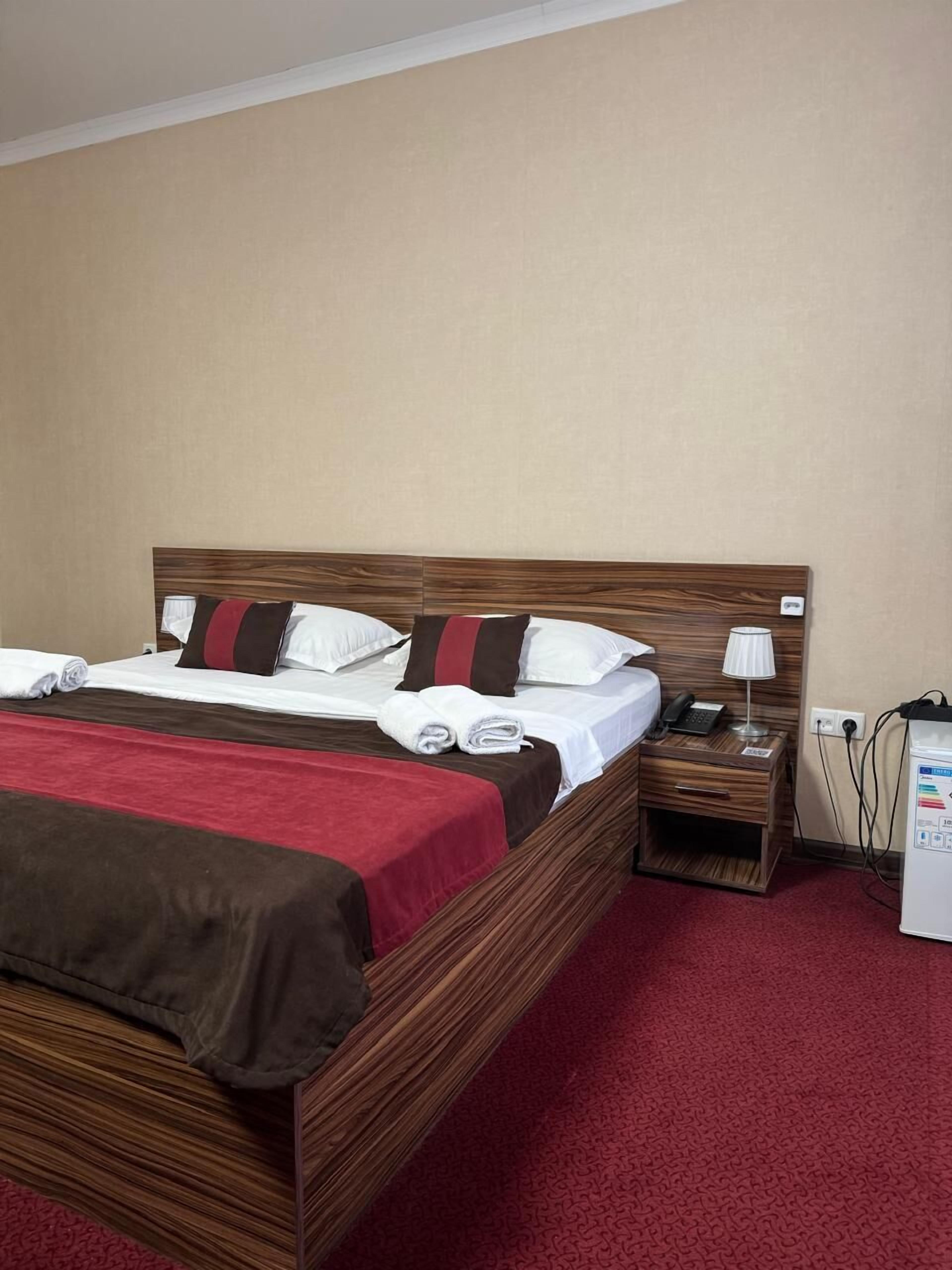 A hotel room with a wooden bed, red and brown bedding, a bedside table with a lamp, and a small refrigerator.