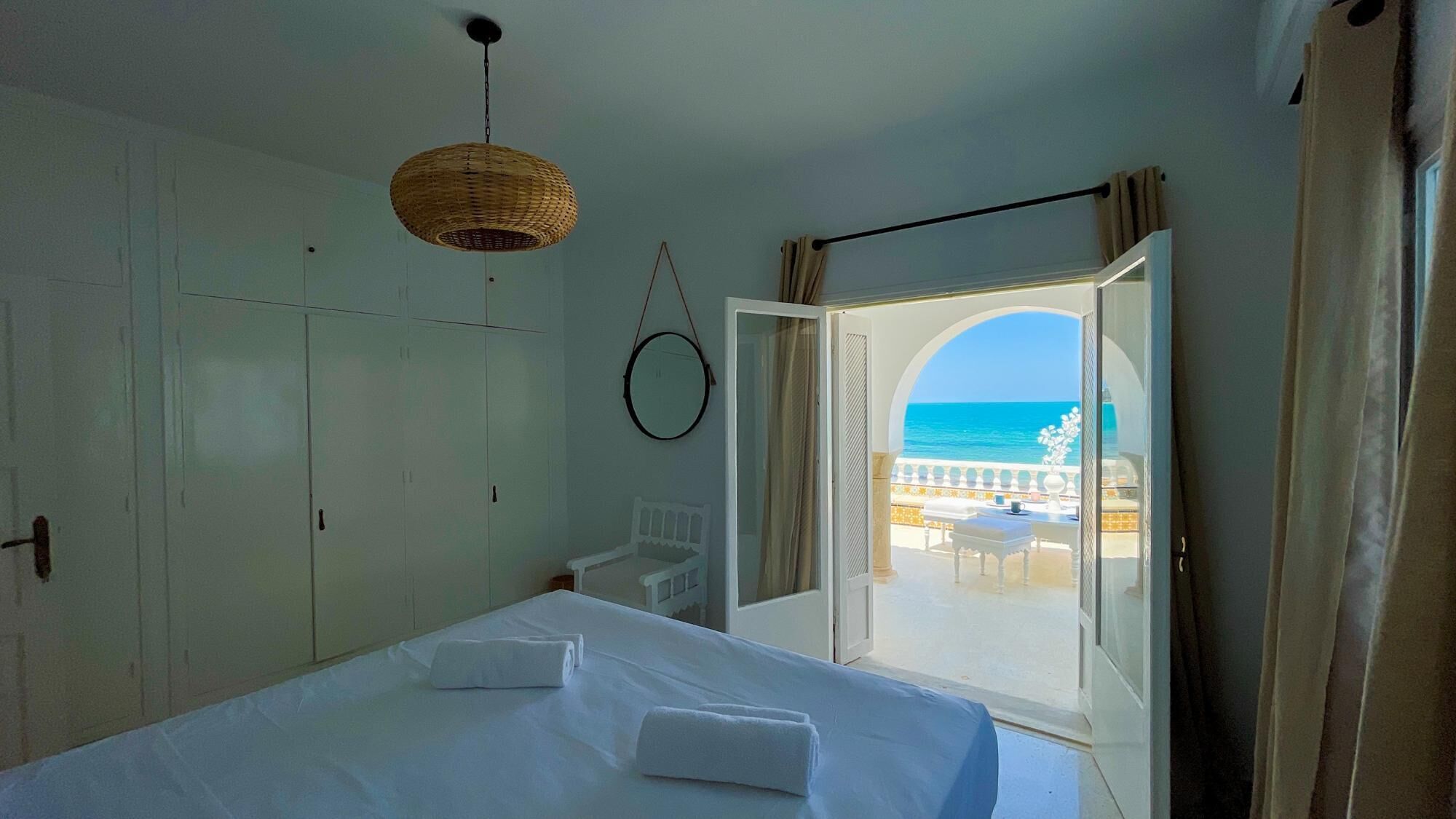 Quarto com cama, luminária pendente, guarda-roupa e vista para o mar através de uma porta aberta.