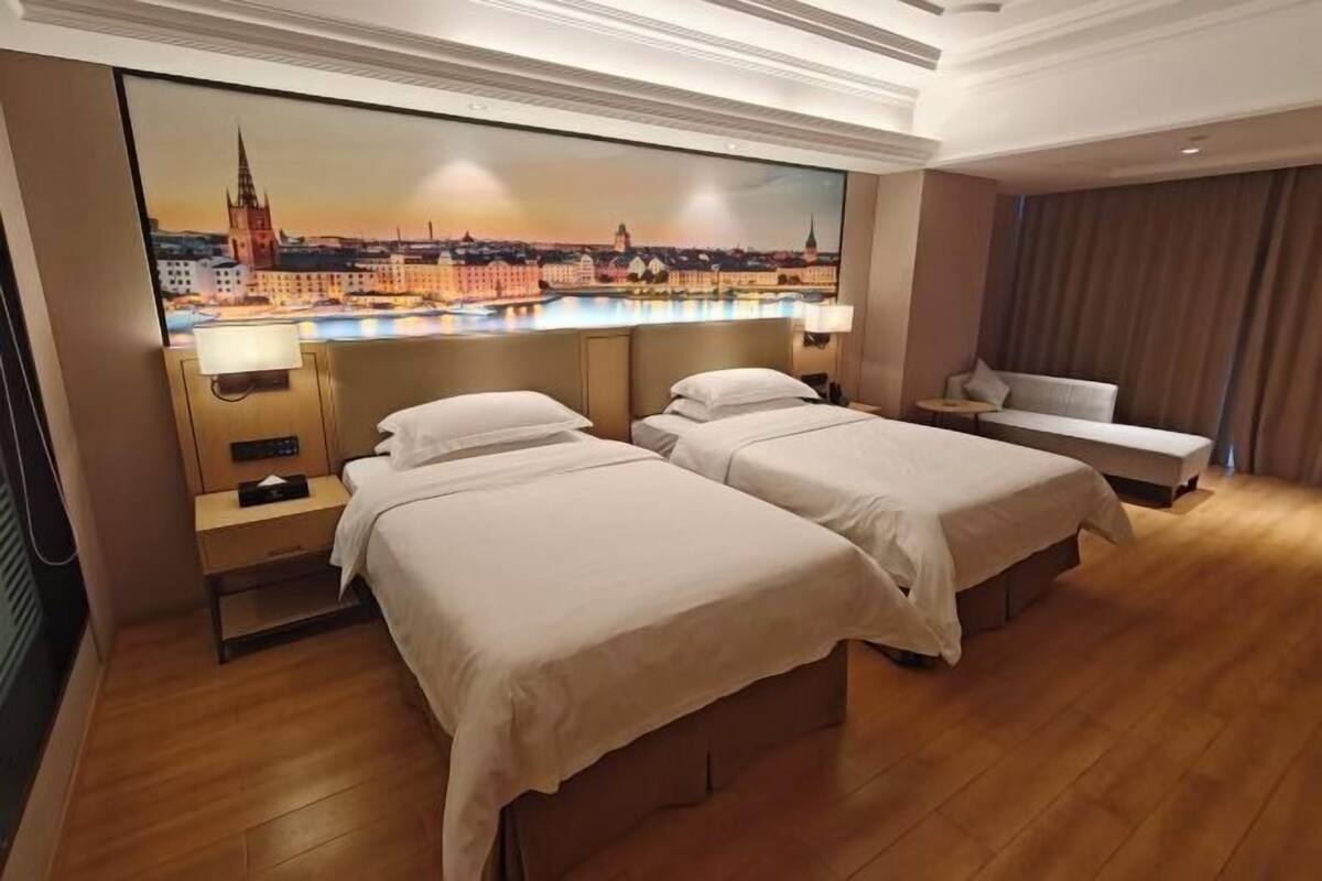 Habitación de hotel con dos camas, un cuadro grande de un paisaje urbano en la pared, suelo de madera y vistas a la ciudad.