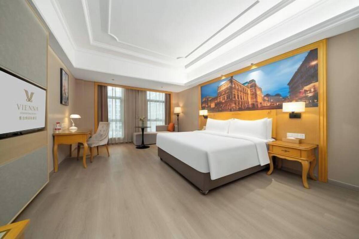 Una habitación de hotel con una cama grande, un escritorio, una silla y un cuadro de un paisaje urbano.