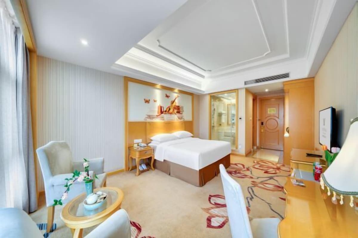 A hotel room with a bed, a desk, a chair, a small table, and a television.