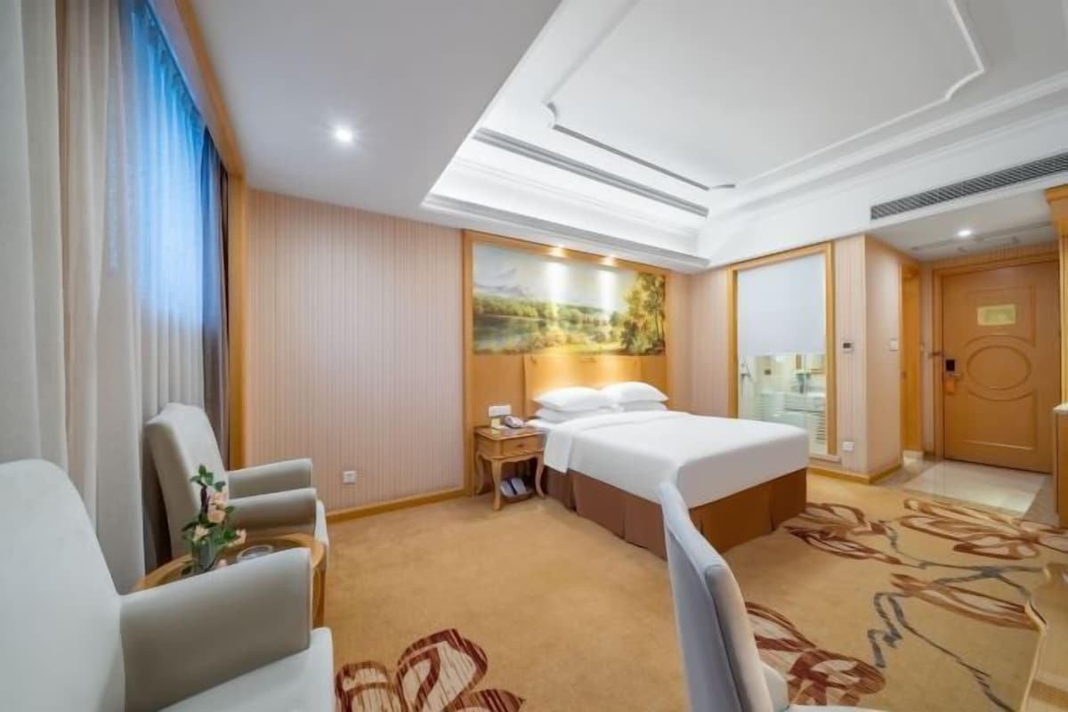 A hotel room with a bed, a seating area with chairs, a small table with a vase, and a painting on the wall.