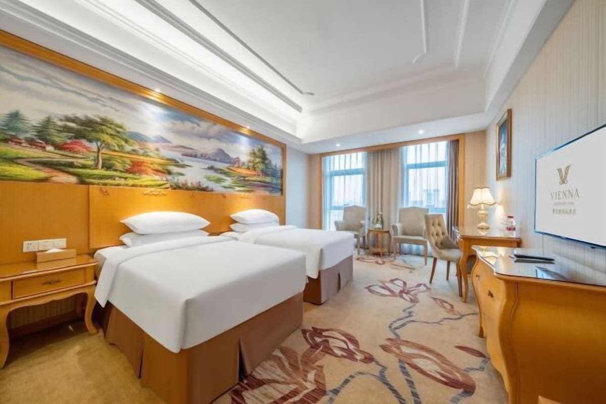 A hotel room with two beds, a desk, a chair, a TV, and a large painting on the wall.