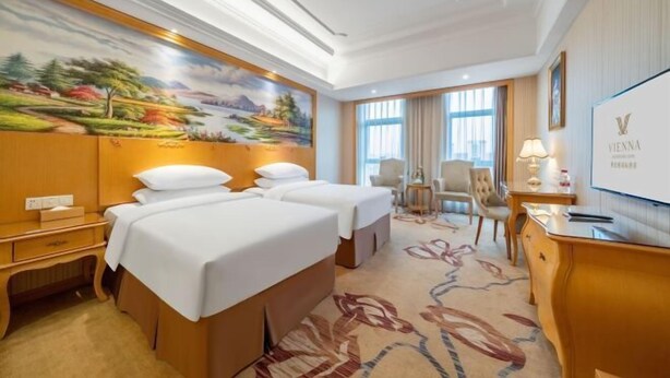 A hotel room with two beds, a desk, a chair, a TV, and a large painting on the wall.