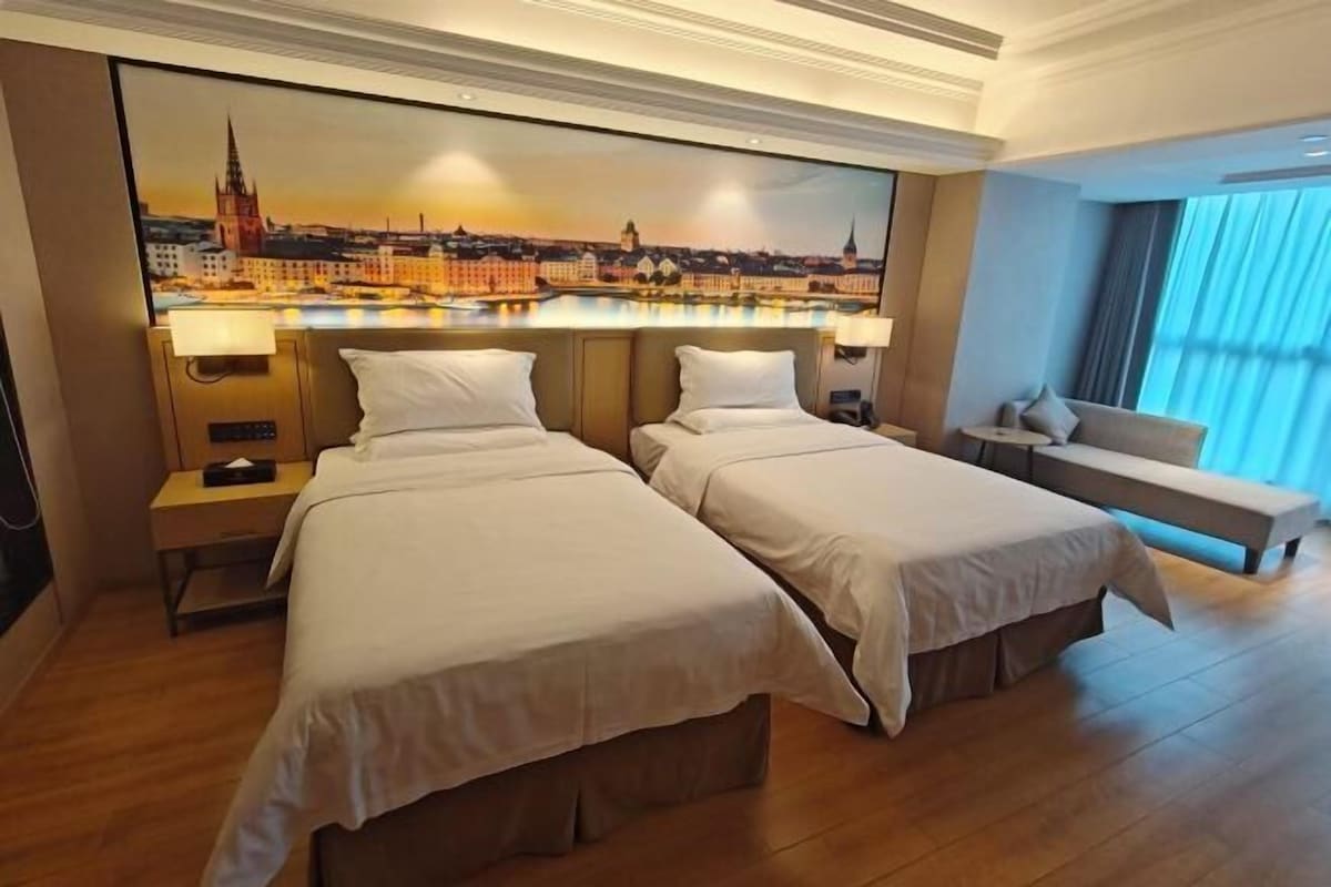 A hotel room with two beds, a bench, a large cityscape painting on the wall, and a window with curtains.