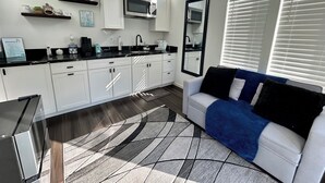 A modern kitchen with white cabinets, a black countertop, and a built-in microwave. There is a large window with blinds, a sofa with a blue throw blanket, and a rug with a geometric pattern. - Cozy, Quaint and Sophisticated Loft Style Tiny Home!  (Flagstaff)